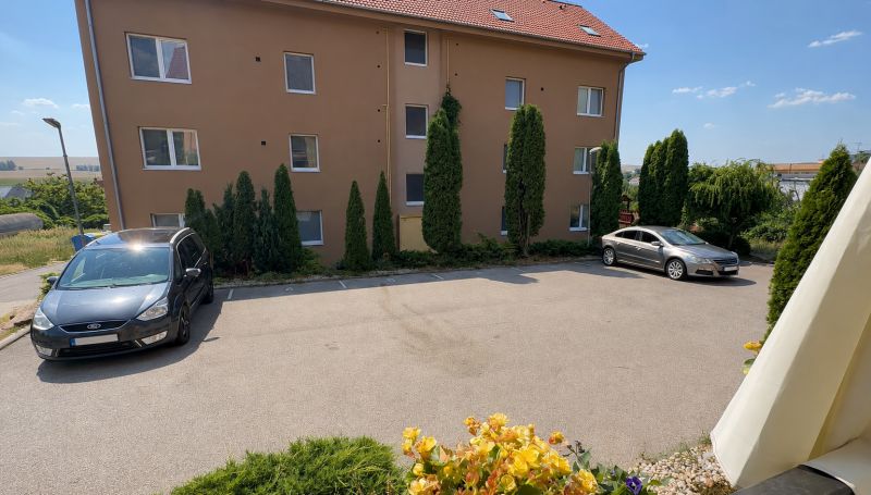 ÚTULNÝ 3-IZBOVÝ BYT V POKOJNOM PROSTREDÍ, LEN KÚSOK OD NITRY - Parkovisko pred bytovkou vo Veľkom Lapáši s autami a zeleňou v okolí.