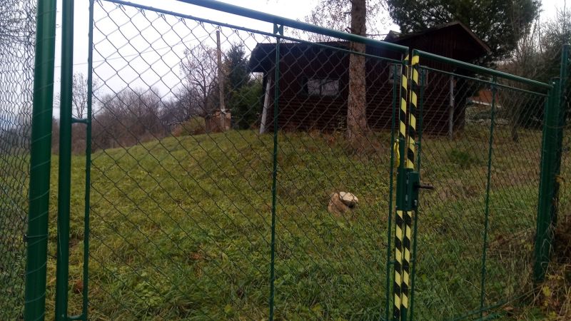 KONSKÉ - pozemok stavebný 1108 m2,okr. Martin  - Brána so zeleným pletivom, vedúca na trávnatý pozemok, blízko drevené obydlie.