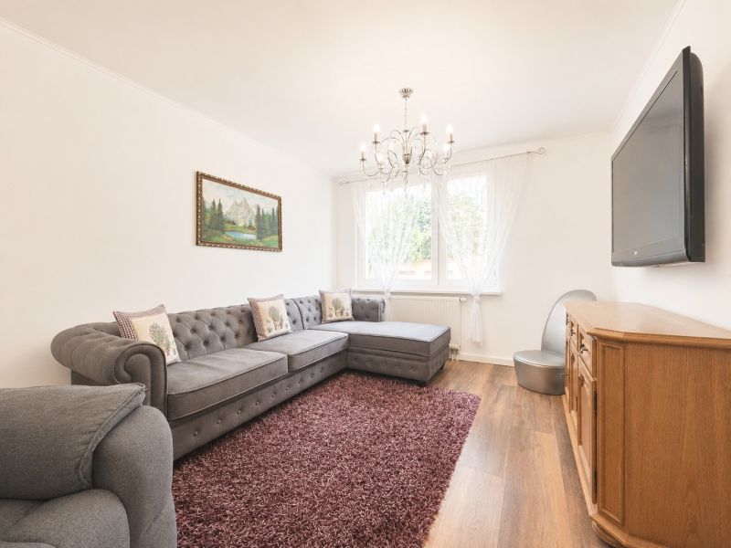 Ponúkame na predaj 4-izbový byt v Šamoríne - Obývačka so sivou sedačkou, lustrom, televízorom a podlahou s dreveným dekorom v 4-izbovom byte.