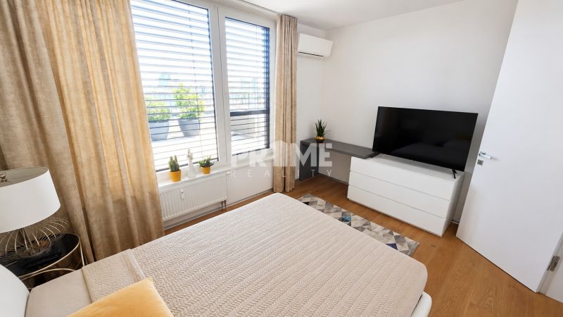PENTHOUSE – moderný 2i byt s výnimočnou 69 m² terasou, AHOJ PARK - Svetlá spálňa s posteľou, televízorom a podlahou s dreveným dekorom v 2-izbovom byte.