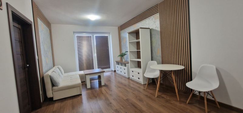 2-IZBOVÝ BYT S VEĽKÝM BALKÓNOM NA PREDAJ - Obývacia izba v 2-izbovom byte so svetlou sedačkou a podlahou s dreveným dekorom.