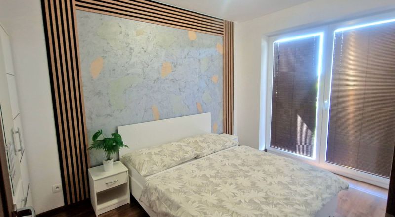 2-IZBOVÝ BYT S VEĽKÝM BALKÓNOM NA PREDAJ - Spálňa v 2-izbovom byte s posteľou, nočným stolíkom a dreveným dekórom na stene.