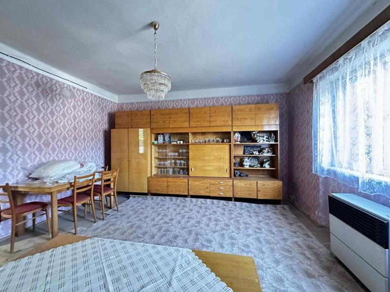 Na predaj starší rodinný dom vhodný na kompletnú prestavbu s  veľkým p - Obývacia izba v rodinnom dome s drevenou skriňou, stolom a kobercom.