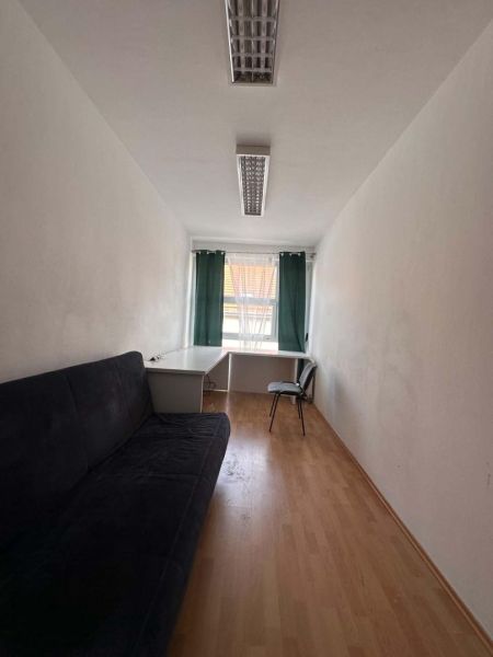 Na prenájom menšia  kancelária v centre mesta DS - 12,94 m2 - Úzka kancelária s pohovkou, stolom a stoličkou; podlaha s dreveným dekorom.