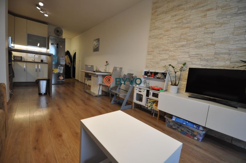 3-IZBOVÝ TEHLOVÝ BYT V DUBNICI NAD VÁHOM | 68 m² | BALKÓN | VLASTNÉ KÚ - Obývacia izba s podlahou s dreveným dekorom, kuchyňou, jedálenským stolom a TV skrinkou.