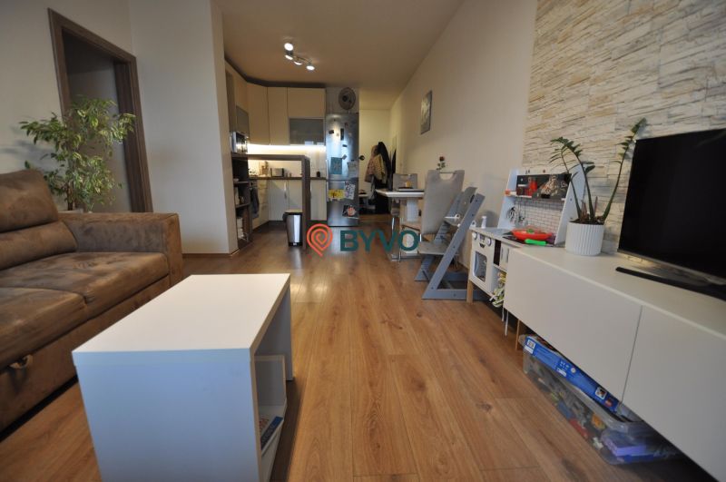 3-IZBOVÝ TEHLOVÝ BYT V DUBNICI NAD VÁHOM | 68 m² | BALKÓN | VLASTNÉ KÚ - Obývacia izba v 3-izbovom byte s podlahou s dreveným dekorom, sedacou súpravou a kuchyňou.