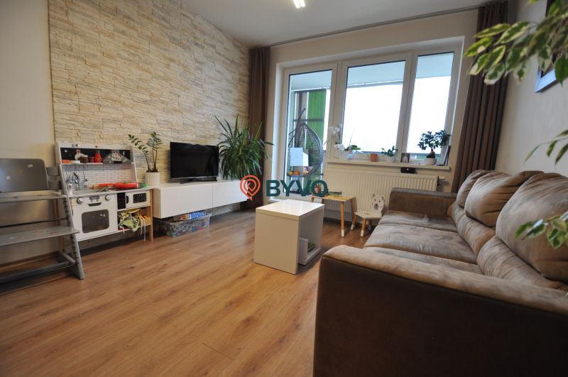 3-IZBOVÝ TEHLOVÝ BYT V DUBNICI NAD VÁHOM | 68 m² | BALKÓN | VLASTNÉ KÚ - Obývačka v 3-izbovom byte s podlahou s dreveným dekorom a kamenným obkladom na stene.