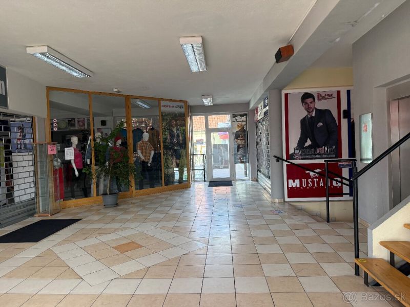 Lukratívny obchodný priestor v Centre Topolčian   - Chodba obchodných priestorov so sklenenými výkladmi, kvetinou a reklamou na stene.
